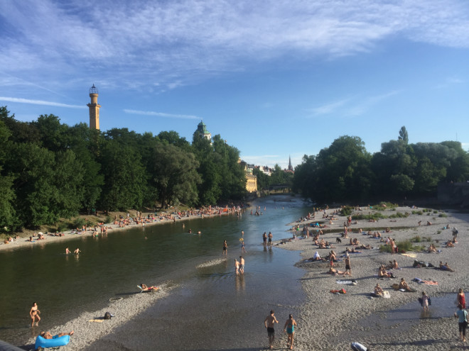 Im Sommer tummeln sich die Münchner auf den Sand/Stein-Bänken der Isar. Das Dach vom Müllerischen Volksbad scheint durch die Bäume.