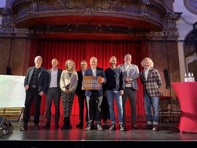 Dr. Christian Scharpf (Mitte, mit Schild) hat heute den Namen "Central Quartier" für das Münchner Bahnhofsviertel verkündet. Mit dabei (v.l.n.r.): Prof. Dr. Klaus Sailer (Hochschule München), Stefan Schillinger (ACCUMULATA), Marie Therese Kröger-Rah (Referat für Arbeit und Wirtschaft), Thomas Linsmayer (Deutsches Theater München), Dr. Christian Scharpf, Benoit Blaser (Bezirksausschuss 2), Reinhard Sigel (Südliches Bahnhofsviertel e.V.) und Prof. Matthias Ottmann (Urban Progress).© Ehret+Klein