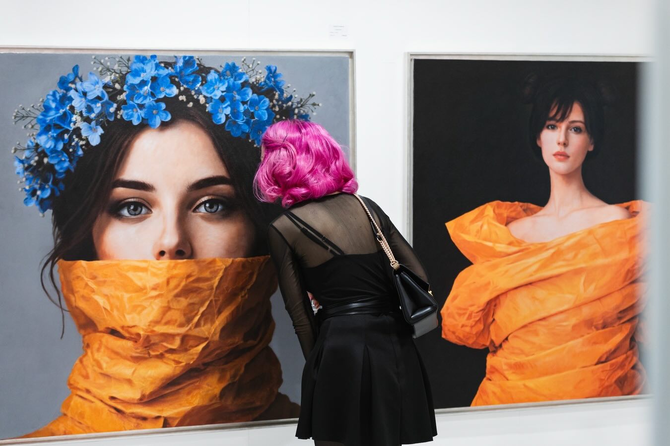 Besucherin mit pinkfarbenen Haaren betrachtet auf der ARTMUC zwei großformatige Frauenporträts mit orangefarbenen Gewändern, eines davon mit blauem Blumenkranz.