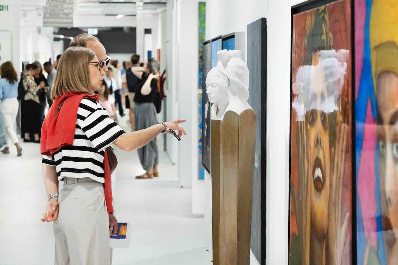 Besucherin mit rot umgelegtem Pullover zeigt auf Kunstwerke neben einer weißen Büste auf der ARTMUC Kunstmesse.