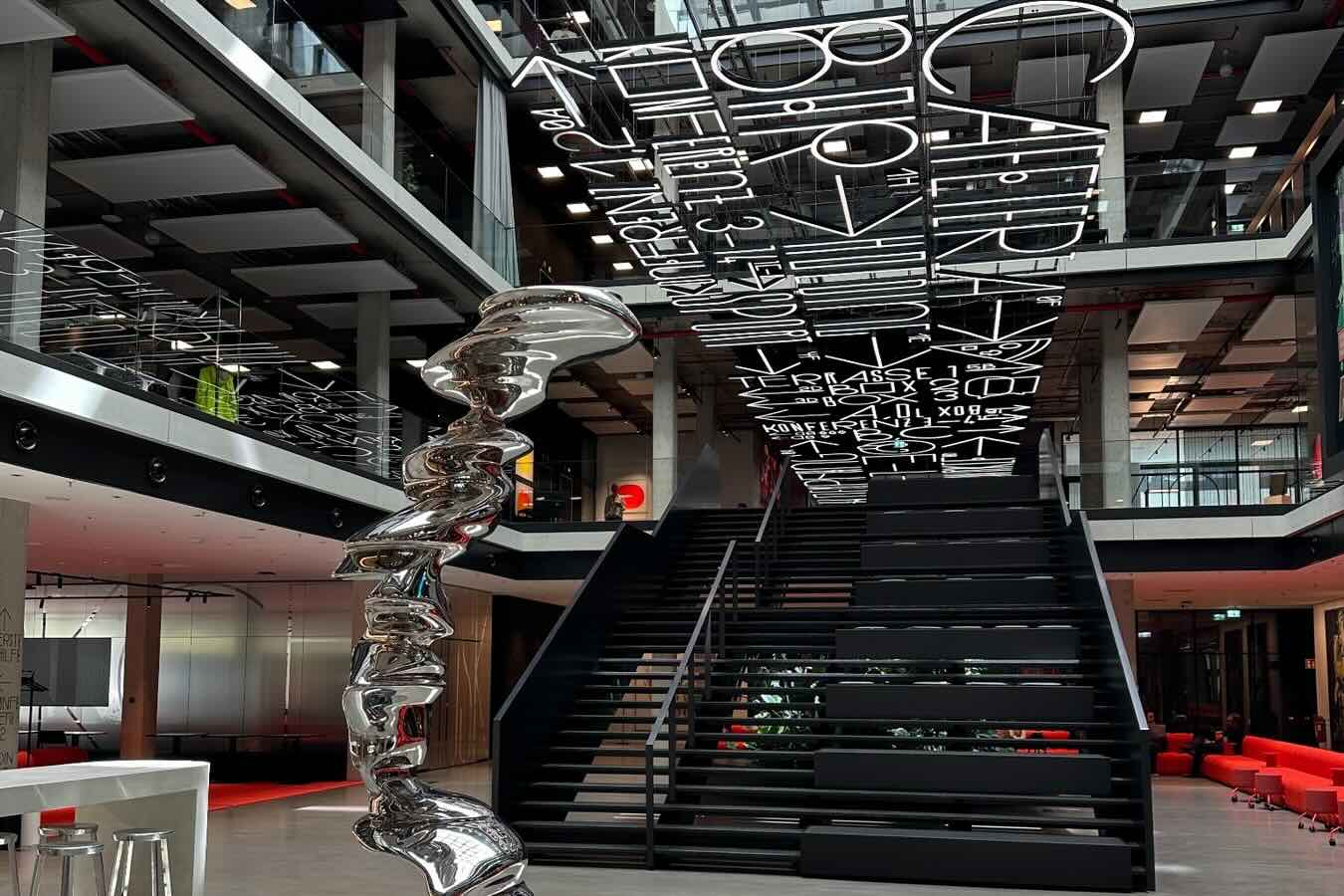 Atrium im House of Communication von Serviceplan mit breiter schwarzer Treppe, leuchtender Schriftinstallation und silberner Skulptur.