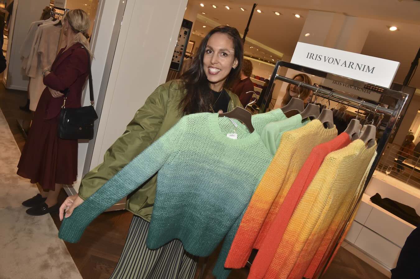 Isabelle Freifrau von Crailsheim hält einen Kaschmirpullover mit mintgrünem Farbverlauf neben weiteren Pullovern in Gelb und Orange bei LODENFREY in München.