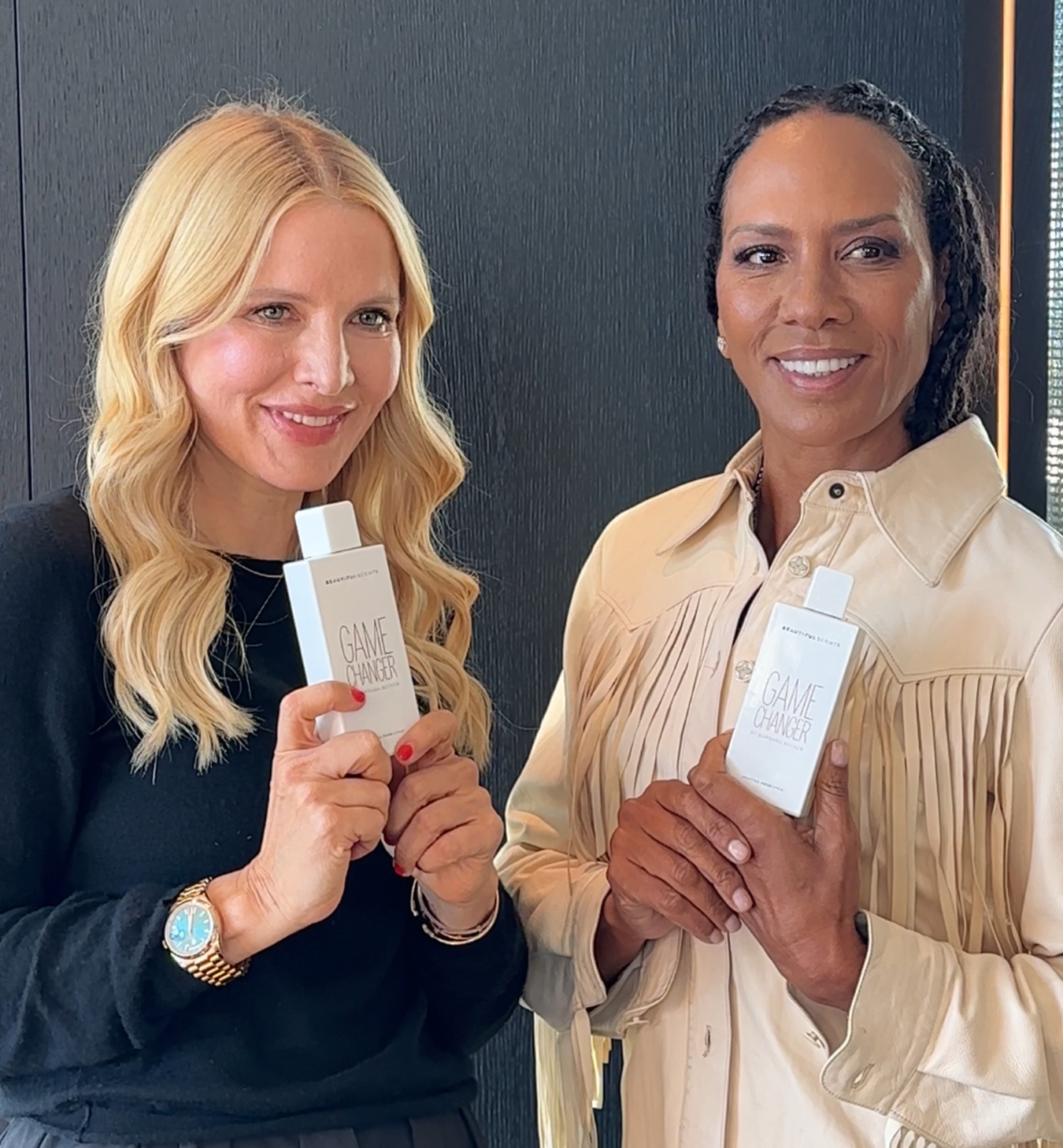 Stefanie Wirnshofer und Barbara Becker lächeln in die Kamera und halten jeweils eine weiße Flasche des „Game Changer“-Sprays von Beautiful Scents in den Händen.