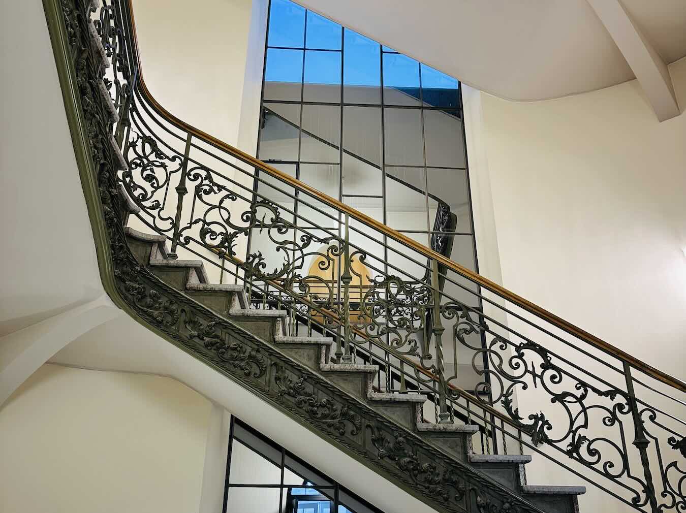 Geschwungene Steintreppe mit reich verziertem Metallgeländer und Holzhandlauf vor großen Fenstern in einem hellen historischen Treppenhaus.