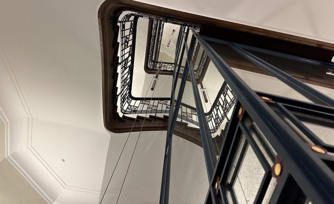 Blick nach oben in ein historisches Treppenhaus mit dunklen Metallgeländern, Holzhandläufen und einem verglasten Aufzug im Treppenauge.